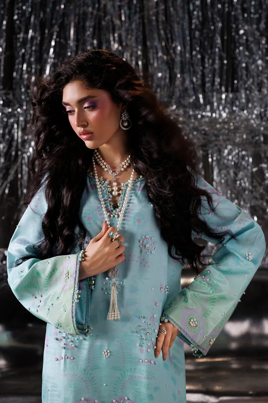 A woman modeling a light blue embroidered traditional outfit with long sleeves, layered pearl necklaces, and statement earrings, posed against a dark silver tinsel backdrop.