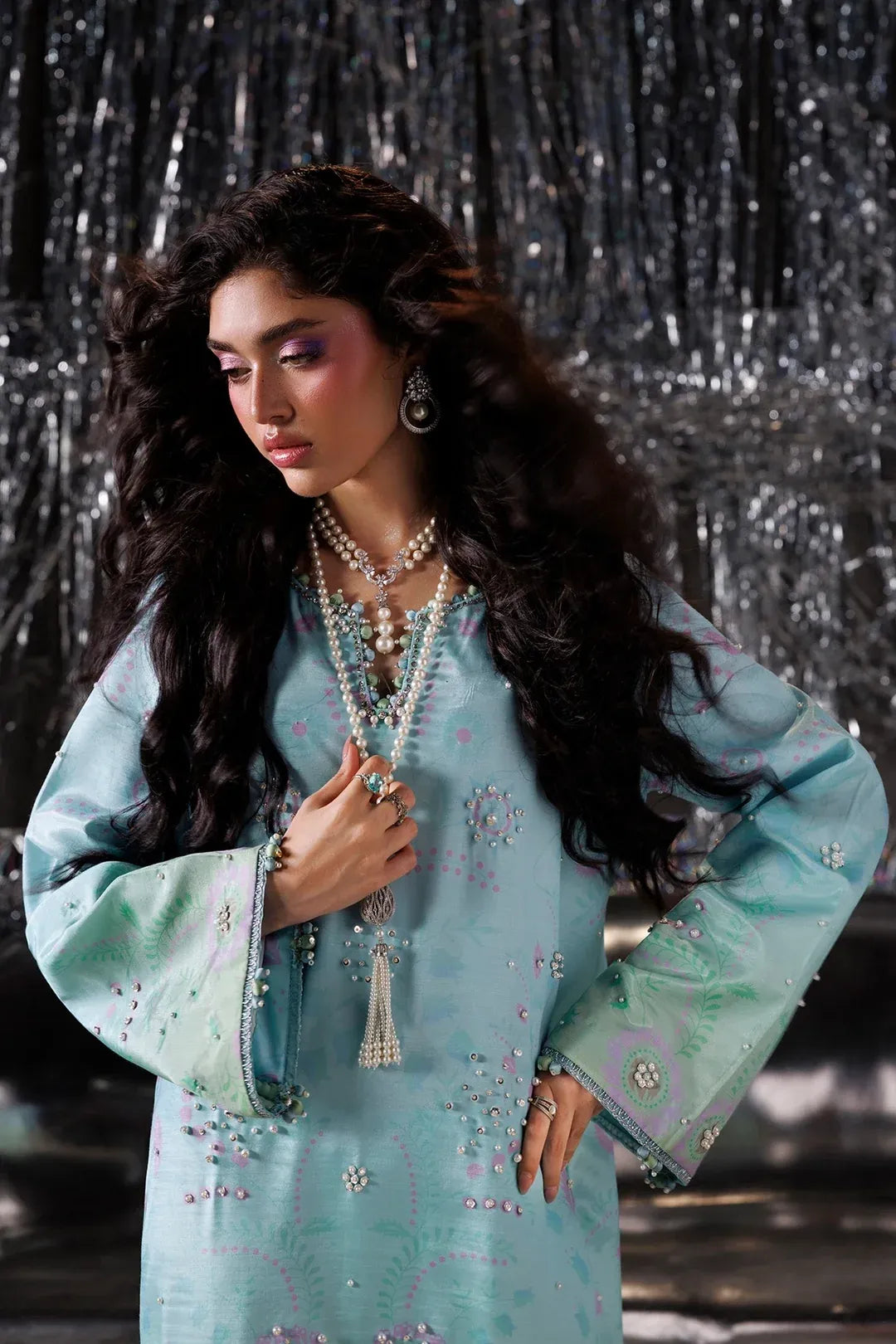 A woman modeling a light blue embroidered traditional outfit with long sleeves, layered pearl necklaces, and statement earrings, posed against a dark silver tinsel backdrop.