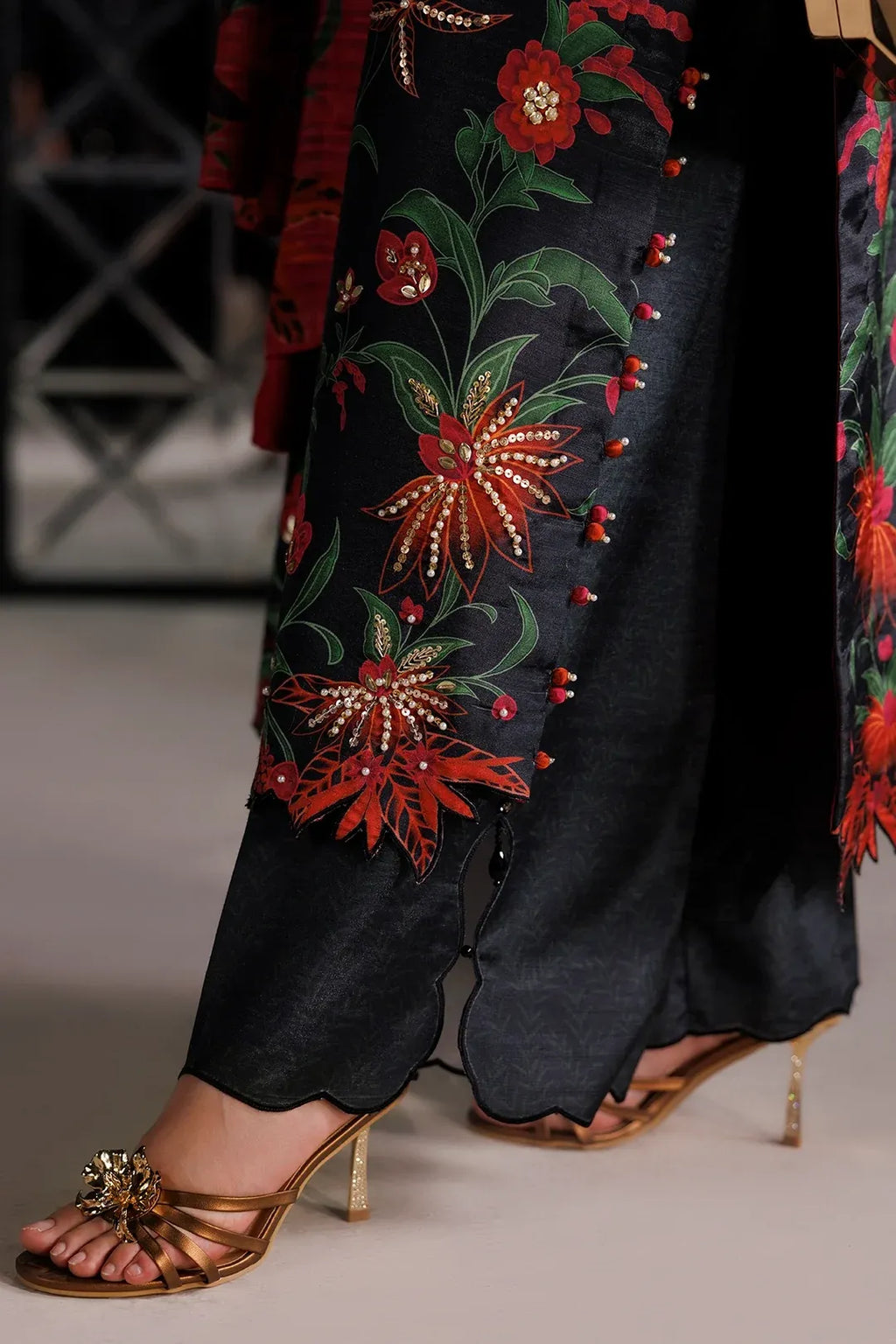 Close-up of a person wearing black wide-leg trousers with large red and green floral embroidery and gold embellishments, paired with gold heeled sandals.