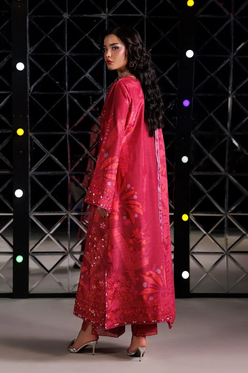 A woman modeling a vibrant pink traditional outfit with floral embroidery and metallic embellishments, standing in front of a dark geometric wall with illuminated dots.