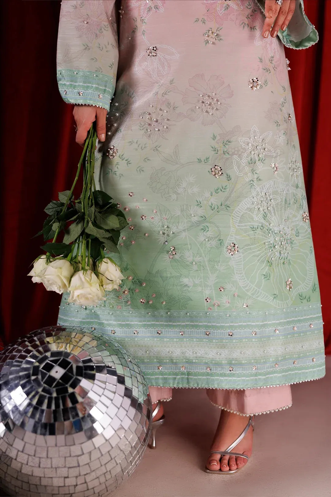 A model wearing a light pink and mint green embroidered traditional outfit, holding a bouquet of white roses next to a large silver disco ball against a red curtain backdrop.