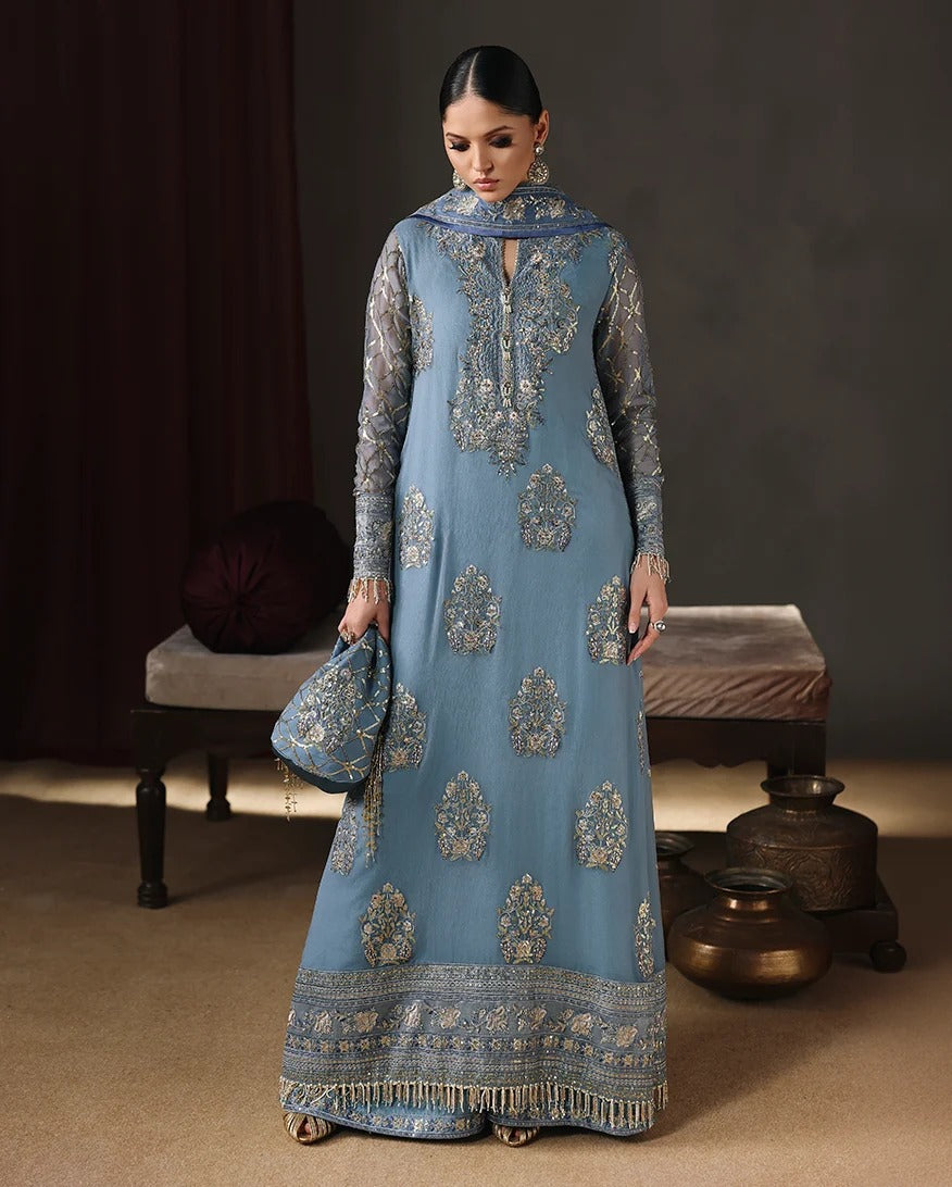 A woman models a traditional blue anarkali gown with intricate gold embroidery, paired with a matching dupatta and a coordinating blue handbag, set against a dark, elegant studio background with vintage props.