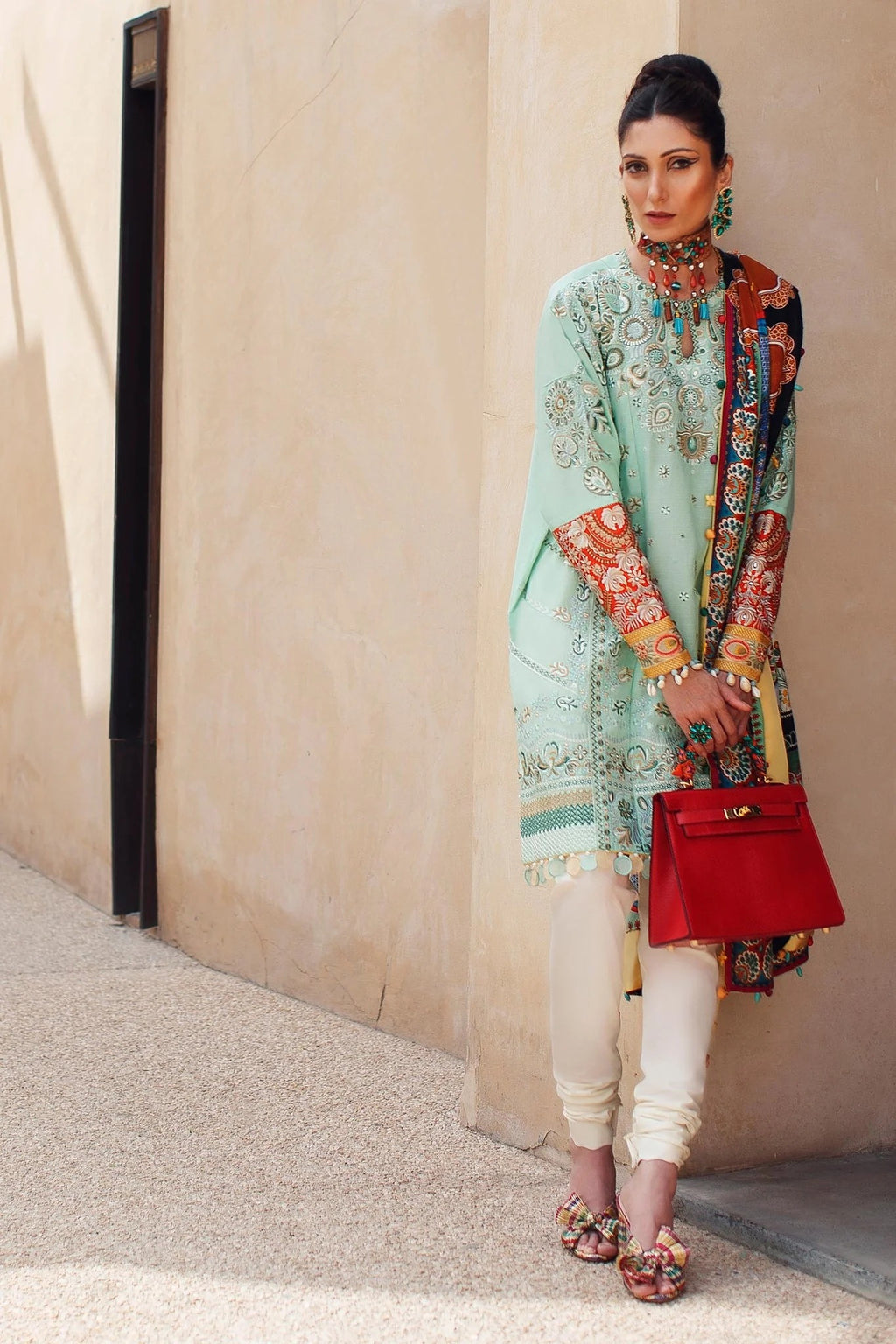 Woman in traditional outfit with a red handbag against a beige wall.