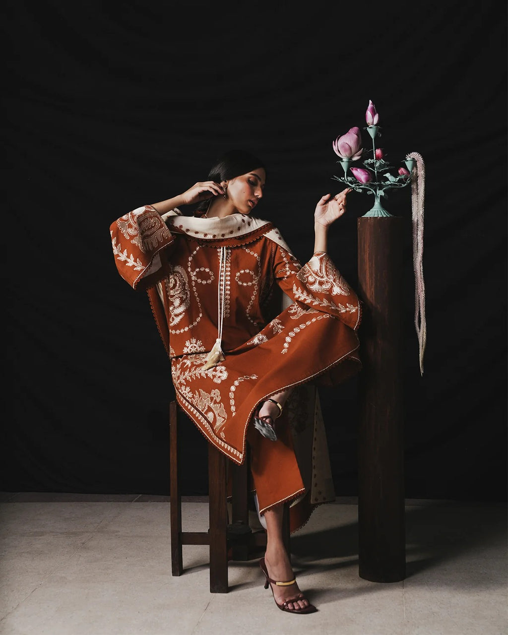 A model wearing a rust-orange caftan with intricate white embroidery and a cream collar, seated on a wooden stool next to a decorative floral stand against a dark studio backdrop.