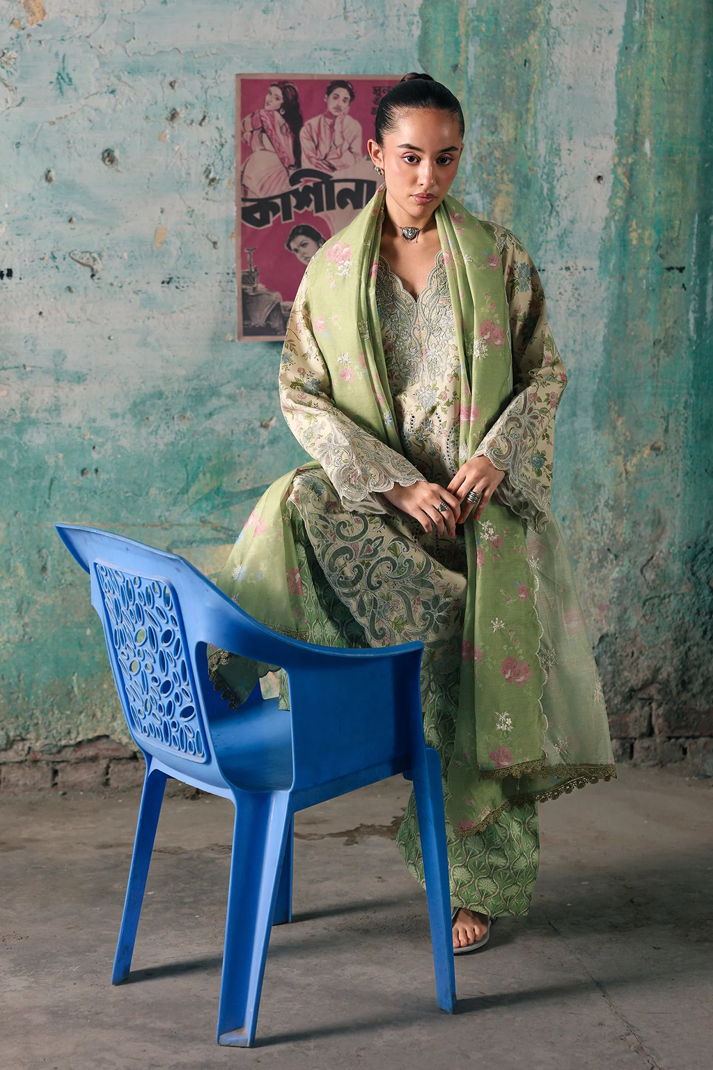 A woman wearing a light green traditional outfit with intricate gold and pink floral embroidery sits on a blue plastic chair against a weathered teal wall. A vintage Hindi poster hangs on the wall behind her.