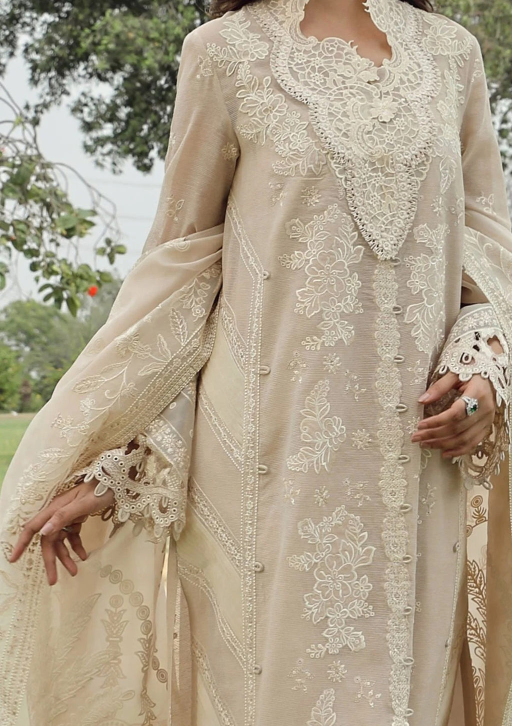 A beige traditional embroidered dress with intricate white floral lace embroidery, shown on a model outdoors with trees in the background.