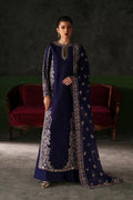 A woman models a deep navy blue traditional outfit with intricate gold embroidery, consisting of a long kurta, matching trousers, and a draped dupatta, set against a dark studio background with red velvet chairs.