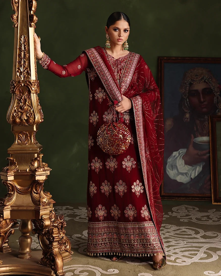 A woman models a deep red embroidered traditional outfit with a matching dupatta and a round embroidered clutch bag, standing next to an ornate gold pillar in front of a dark green wall.
