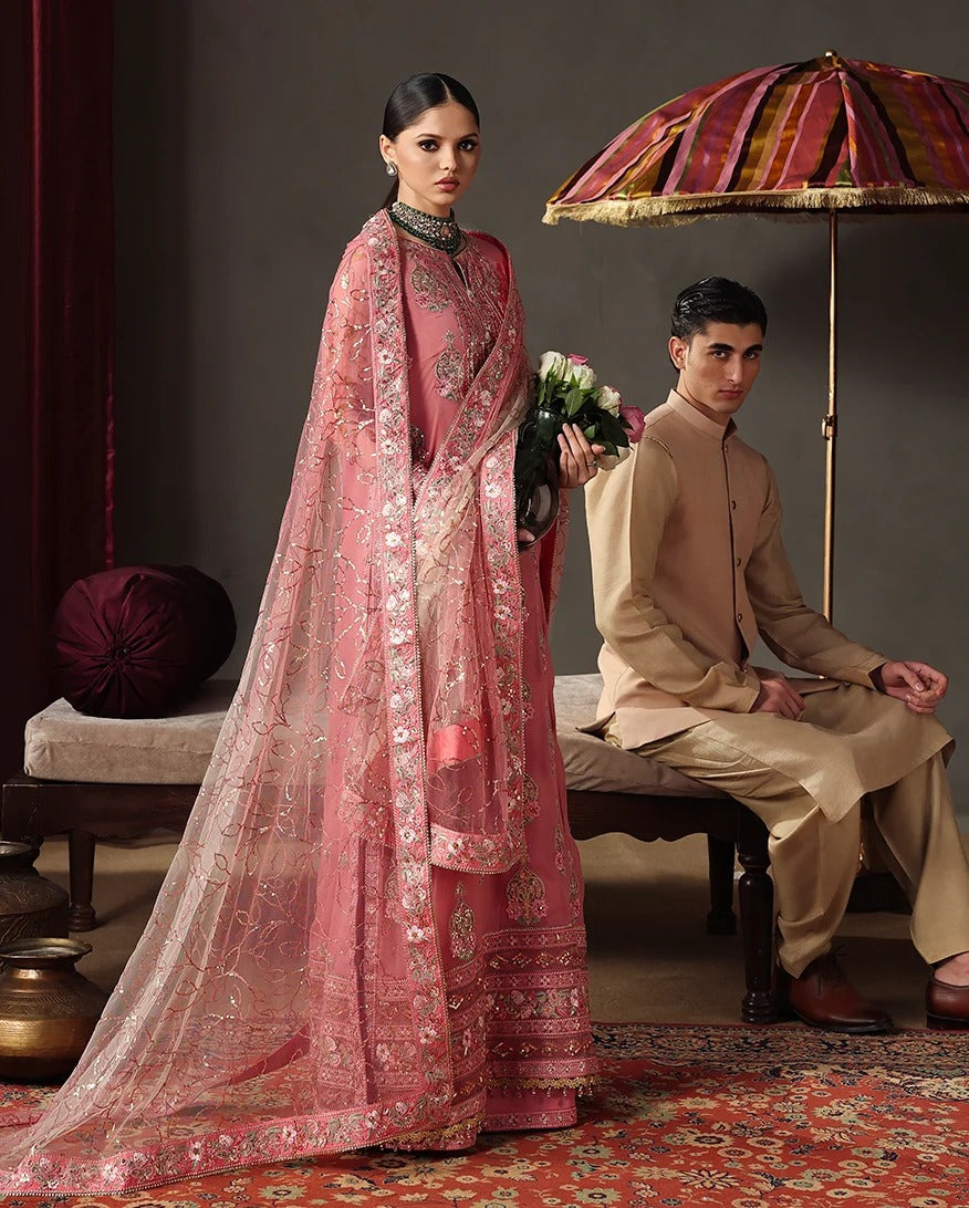 A woman in a pink heavily embroidered traditional outfit with a matching sheer dupatta stands beside a seated man in a beige kurta and trousers under a patterned umbrella in a styled indoor setting.