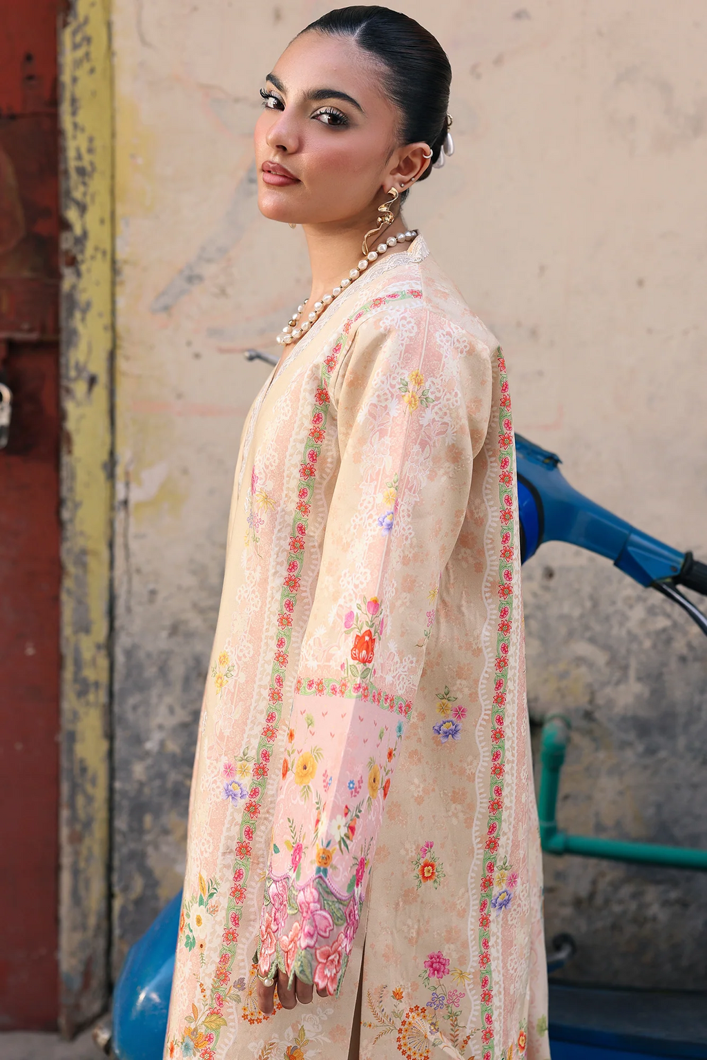 A woman wearing a cream-colored embroidered traditional outfit with floral patterns and a matching jacket, standing in front of a textured wall next to a blue scooter.
