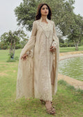A woman wearing a beige embroidered traditional outfit with a long draped jacket and matching pants, standing outdoors near a pond with trees in the background.