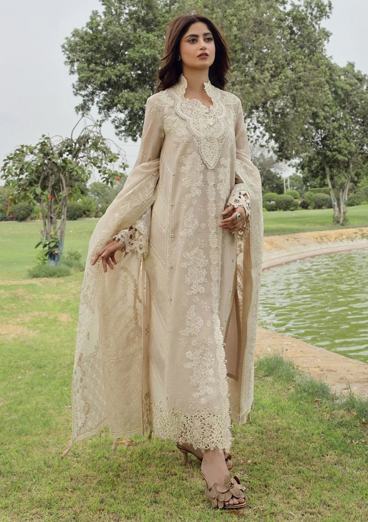 A woman wearing a beige embroidered traditional outfit with a long draped jacket and matching pants, standing outdoors near a pond with trees in the background.
