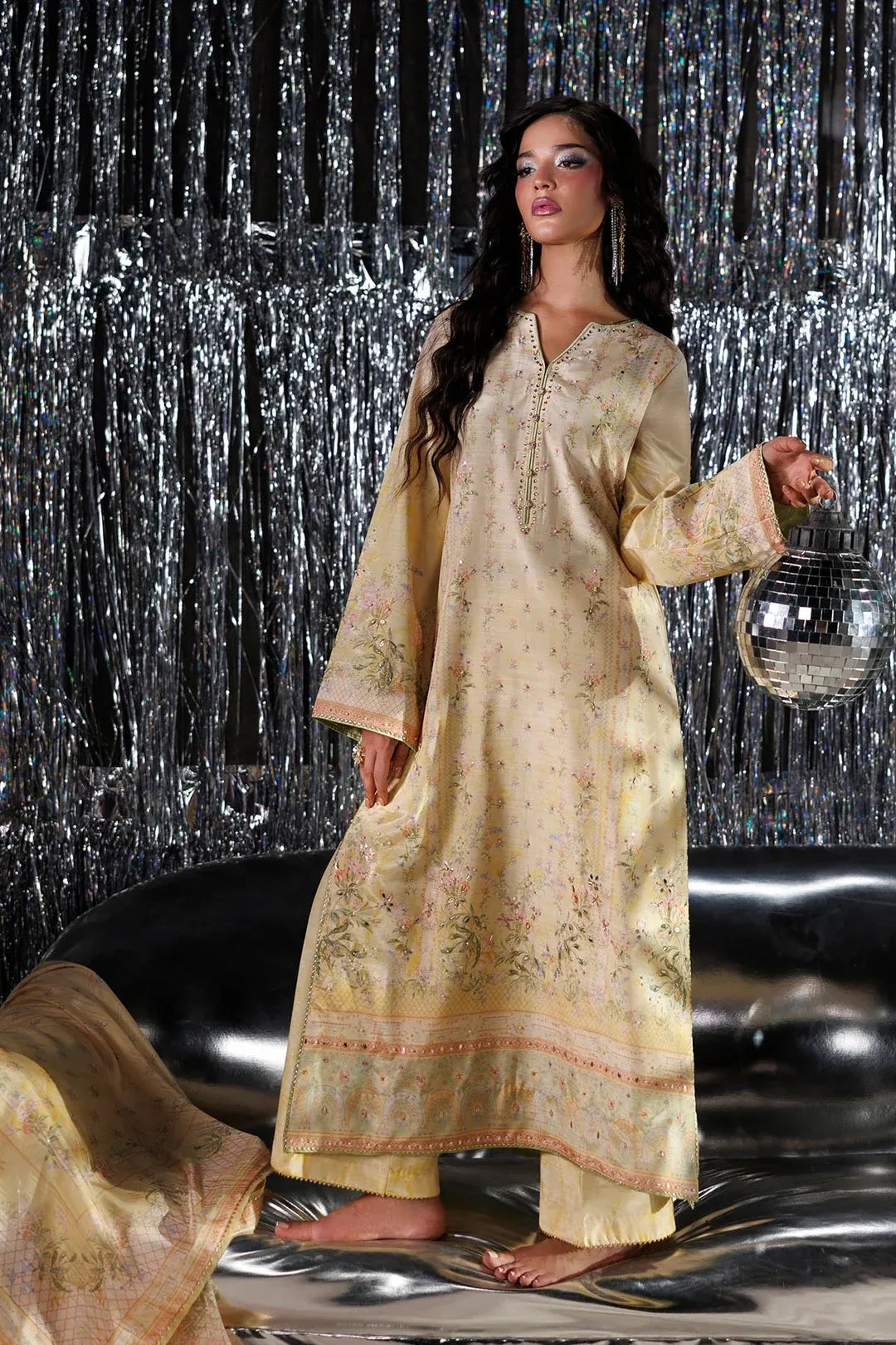 A woman models a traditional long kurta in a pale gold-beige color with delicate floral embroidery and printed motifs, paired with matching trousers. She stands on a reflective silver floor in front of a silver tinsel curtain backdrop, holding a small silver disco ball prop.