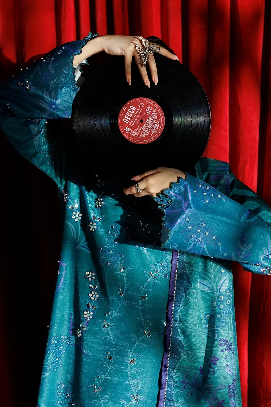 A person in a teal embroidered robe holds a black vinyl record in front of their face, with a red curtain backdrop.