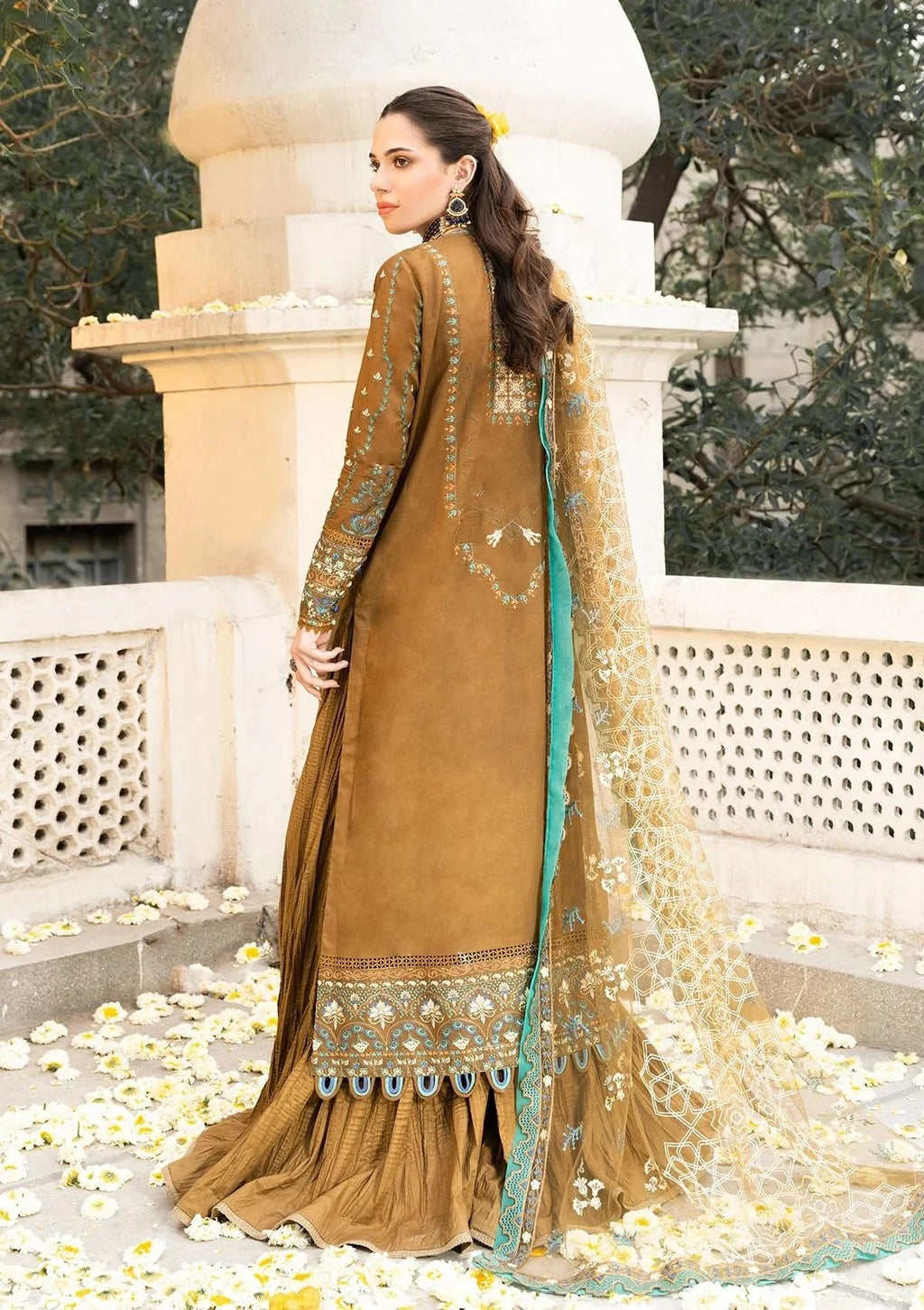 A woman modeling a traditional brown salwar kameez suit with intricate gold embroidery and a teal-green border, paired with a matching sheer dupatta, set against a stone architectural background with scattered white flower petals.