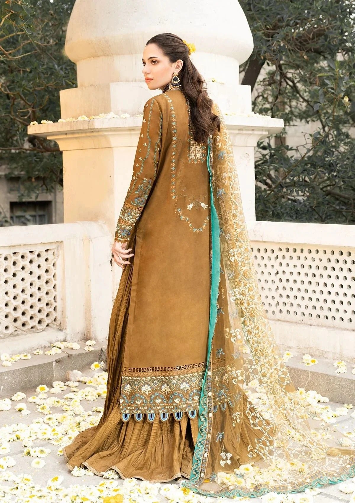 A woman modeling a traditional brown salwar kameez suit with intricate gold embroidery and a teal-green border, paired with a matching sheer dupatta, set against a stone architectural background with scattered white flower petals.