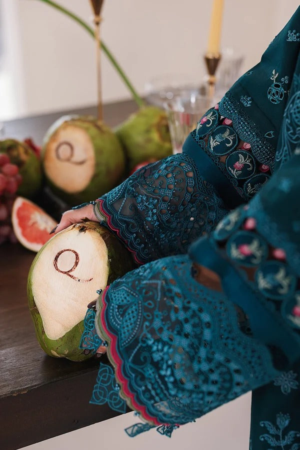 Person holding a fruit with a letter 'Q' on it, wearing a teal embroidered garment.
