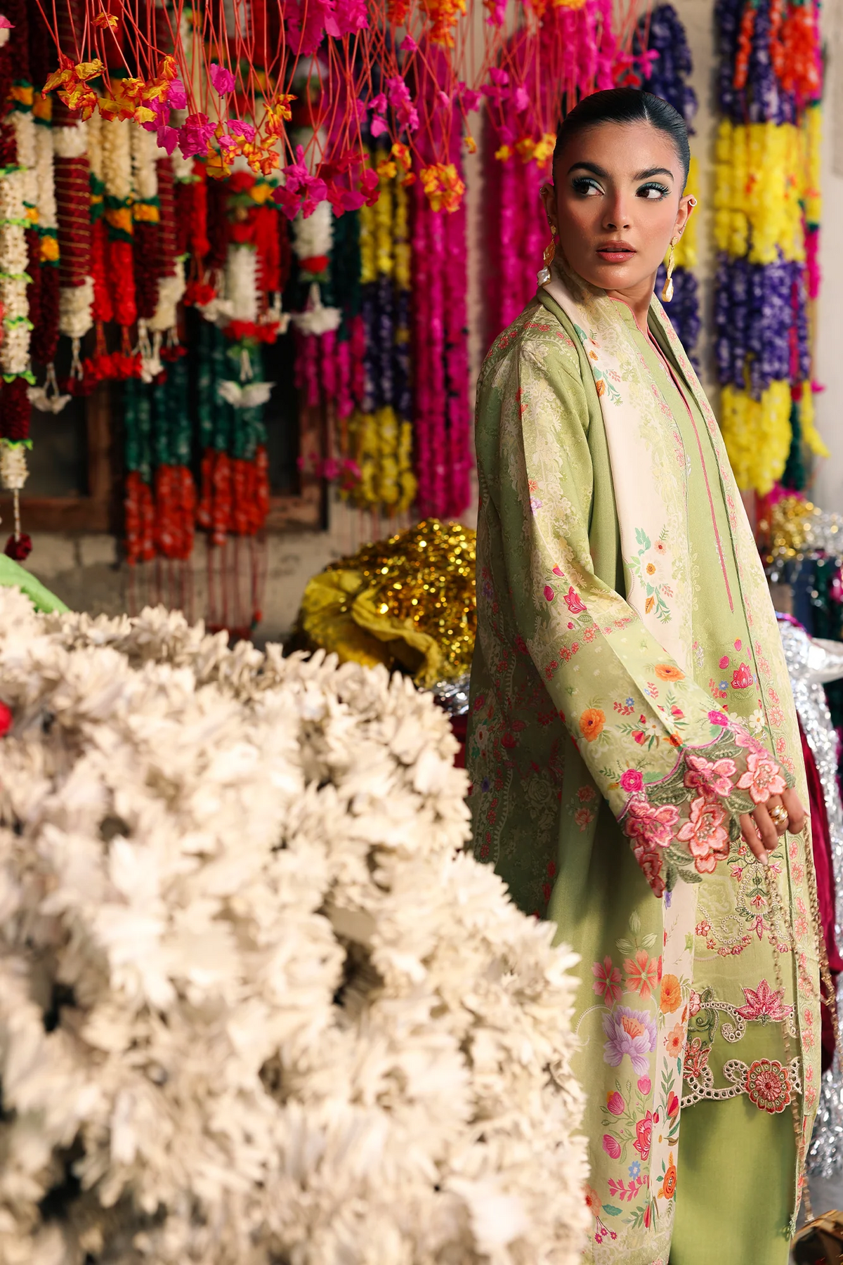 A woman models a light green embroidered kurta with floral patterns in a vibrant, colorful shop setting filled with hanging flower garlands.