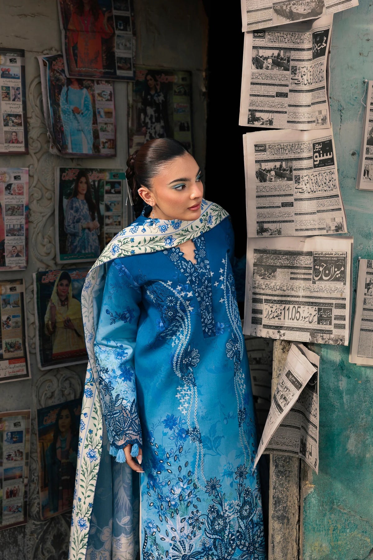 A woman modeling a blue embroidered traditional outfit with a matching patterned dupatta, standing in a doorway surrounded by posters and newspaper clippings.
