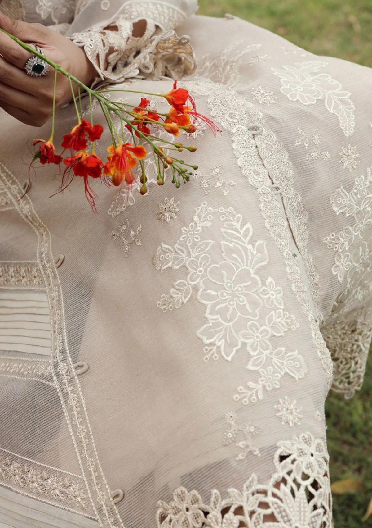 A close-up of a sheer beige lace garment with white floral embroidery and lace trim, held by a hand with a ring while touching small red-orange flowers outdoors.