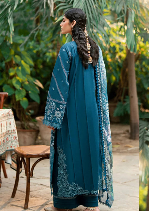 A woman wearing a teal blue traditional outfit with intricate embroidery, shown from the back in an outdoor garden setting.