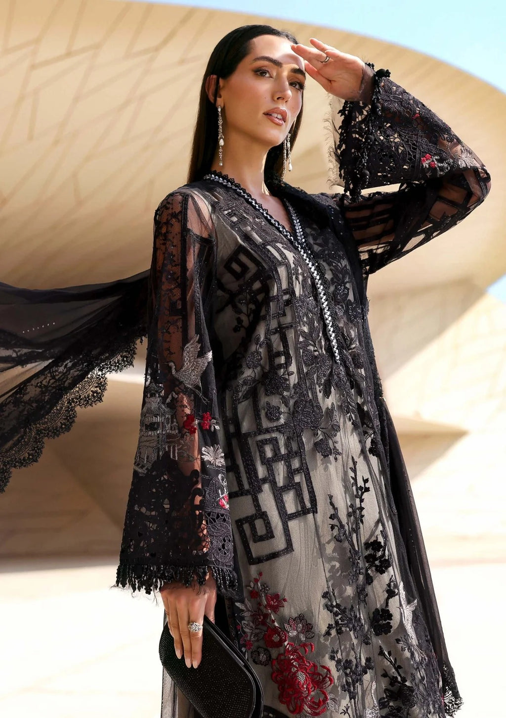 A woman wearing a black sheer embroidered tunic with long sleeves and red floral embroidery, standing outdoors in front of a beige architectural structure.