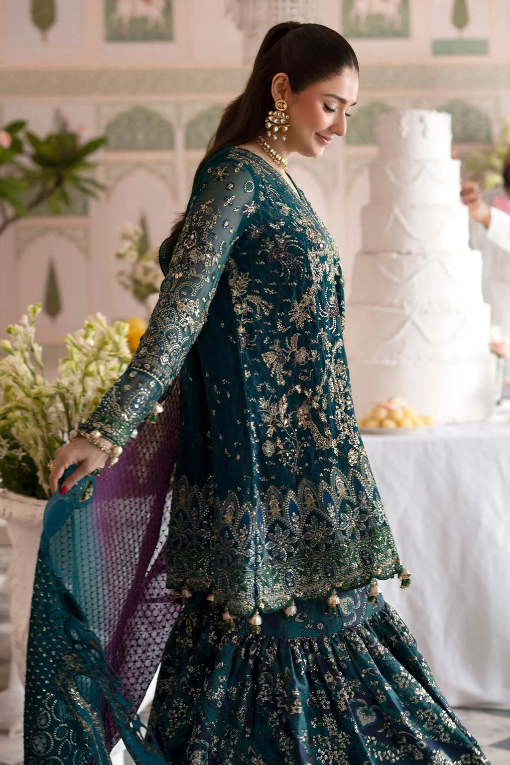 A woman wearing an ornate teal blue and gold embroidered traditional outfit with a matching dupatta, photographed in an indoor event setting with a decorated cake in the background.