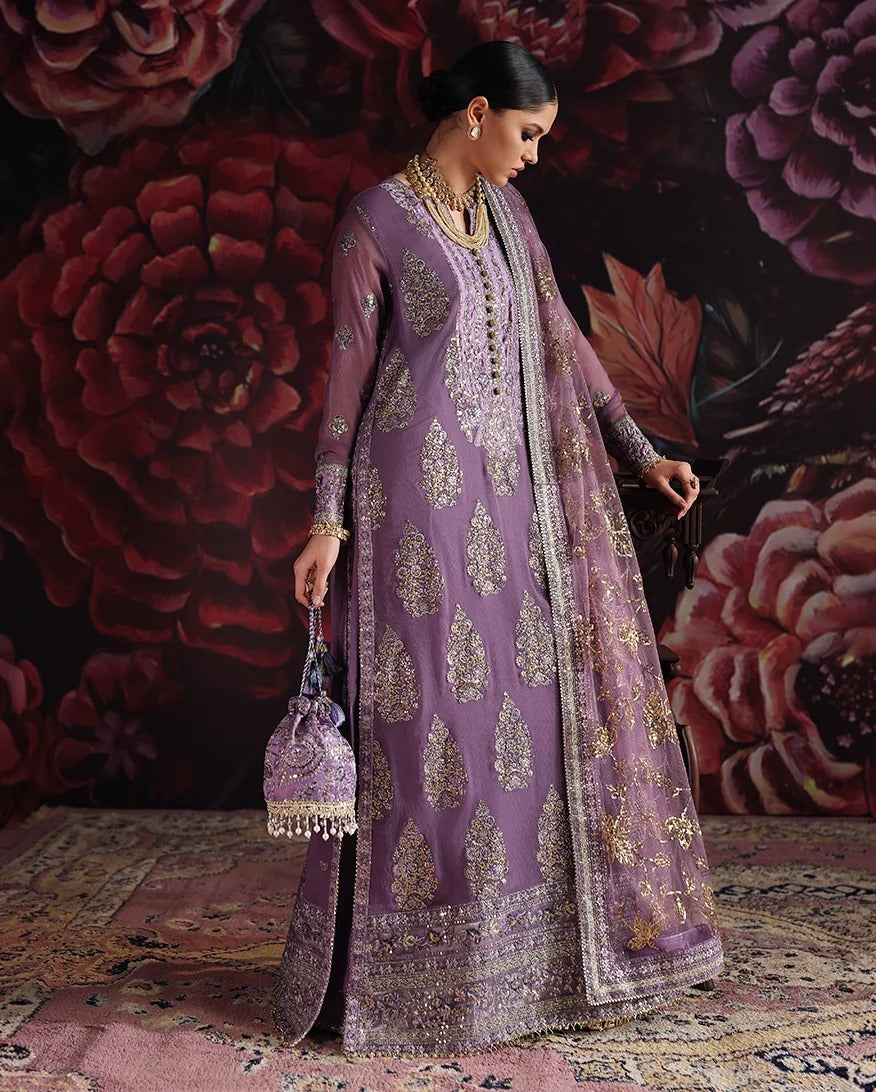 A woman models a lavender purple Anarkali suit with intricate gold and silver embroidery, holding a matching embroidered purple clutch bag against a dark floral wall backdrop.