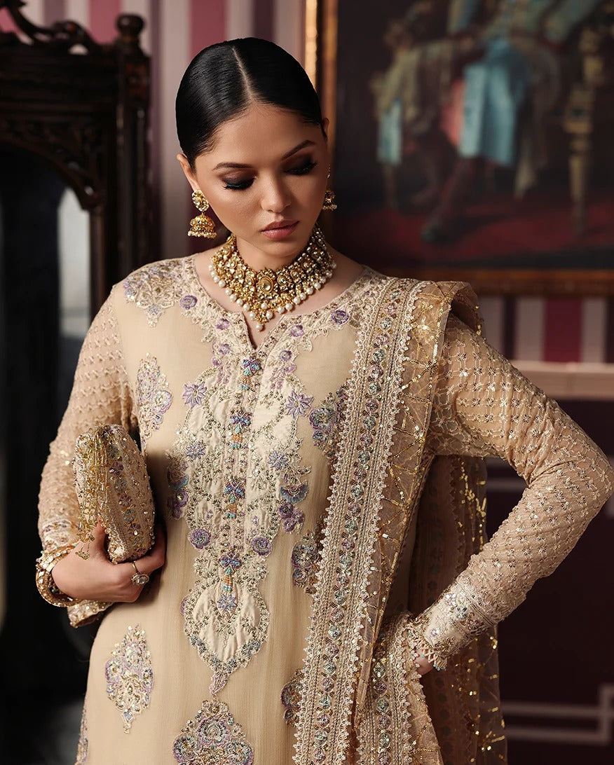 A woman models a traditional beige embroidered kurta set with a matching sheer dupatta and gold jewelry in an indoor setting with a painting in the background.