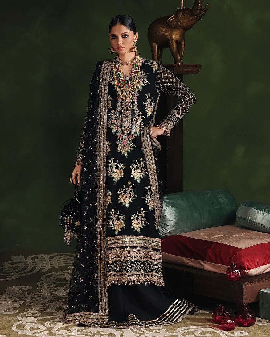 A woman models a traditional black embroidered Anarkali suit with a matching dupatta in a styled studio setting with a dark green background and a wooden bench with cushions.
