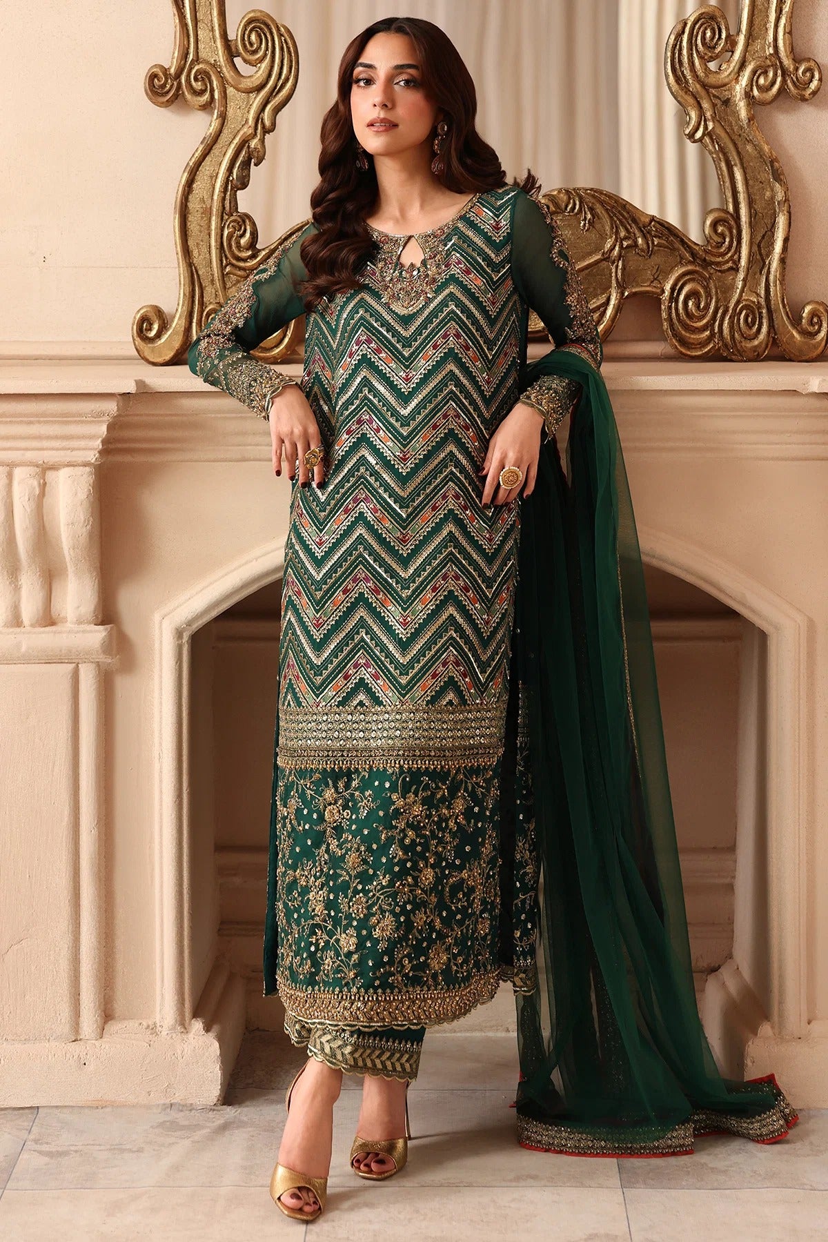 A woman models a traditional Indian ethnic outfit consisting of a deep green kurta with intricate gold and beige embroidery, matching trousers, and a sheer green dupatta, posed in front of a classical fireplace.