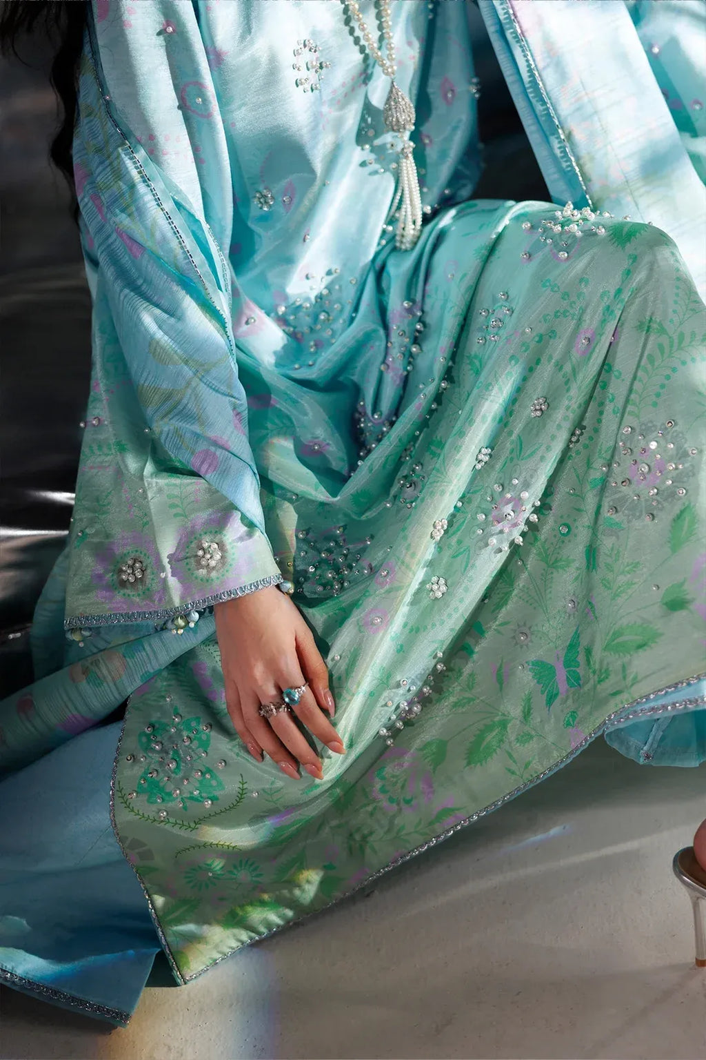A close-up of a person wearing a light blue and mint green embroidered traditional outfit with floral patterns and beadwork, seated indoors.