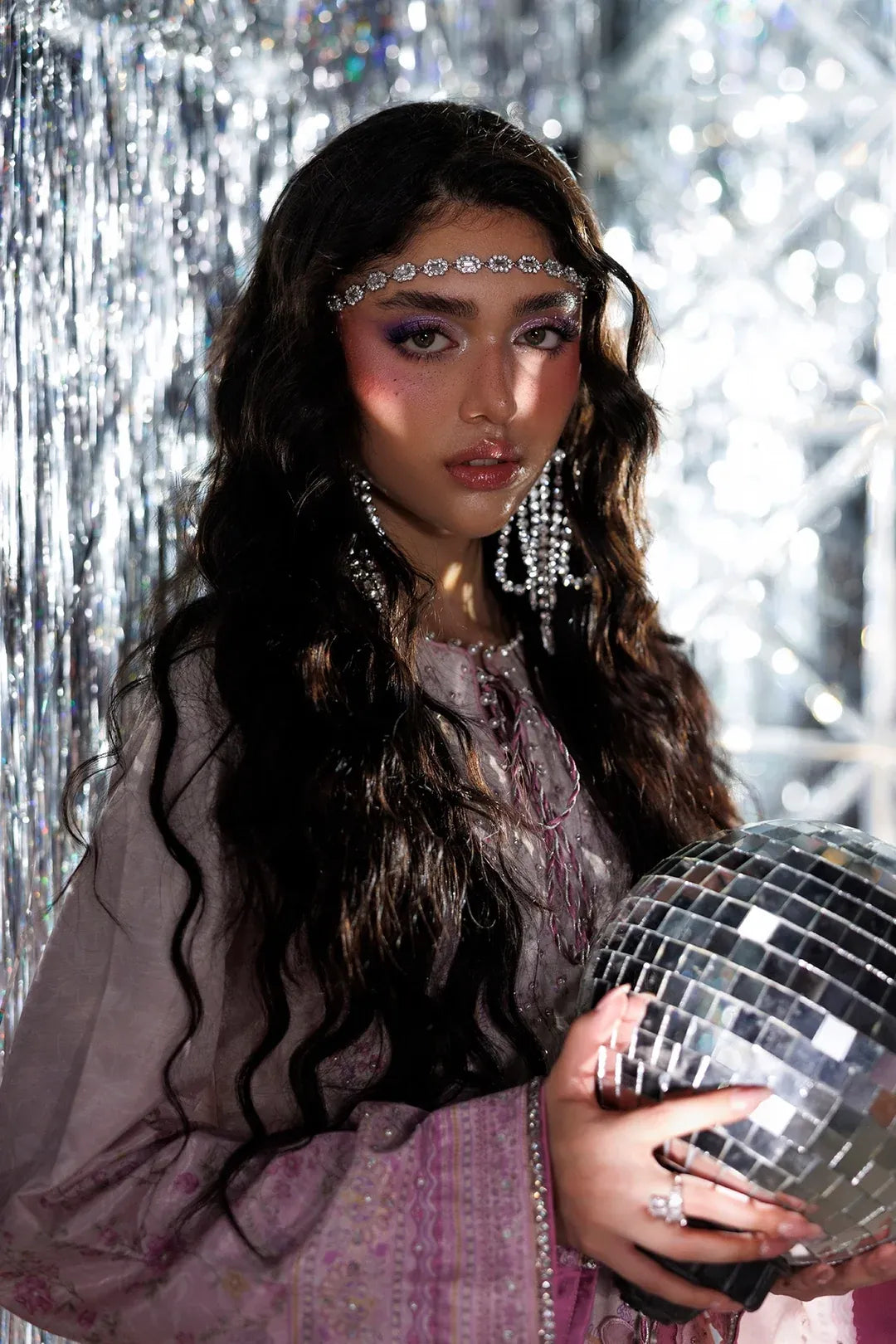 A woman with long dark wavy hair wears a silver rhinestone headband and dangling silver earrings, holding a mirrored disco ball against a silver tinsel backdrop.