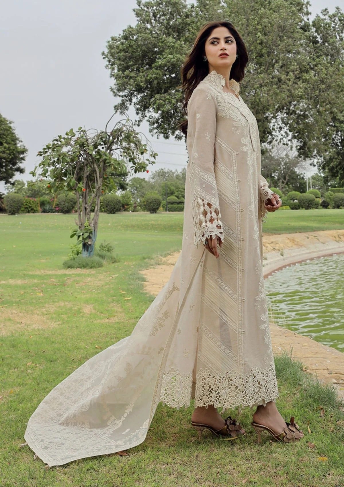 A woman stands outdoors on a grassy lawn beside a pond, wearing a long cream-colored embroidered dress with a matching dupatta and brown heeled sandals.