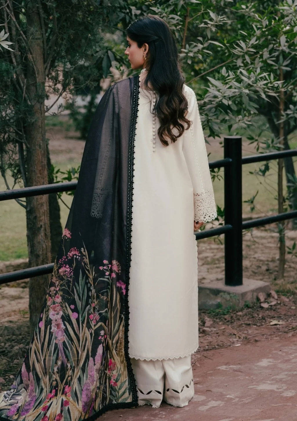 A woman stands outdoors wearing a cream-colored traditional outfit with a black dupatta featuring a pink and green floral print. She is photographed from the back against a background of trees and a fence.