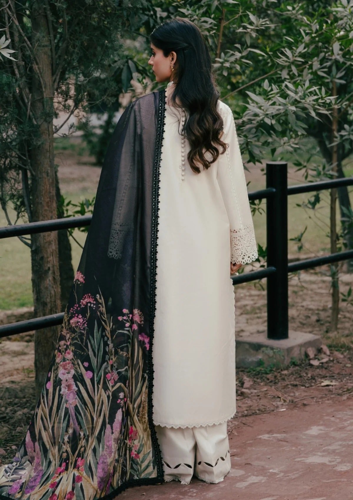 A woman stands outdoors wearing a cream-colored traditional outfit with a black dupatta featuring a pink and green floral print. She is photographed from the back against a background of trees and a fence.