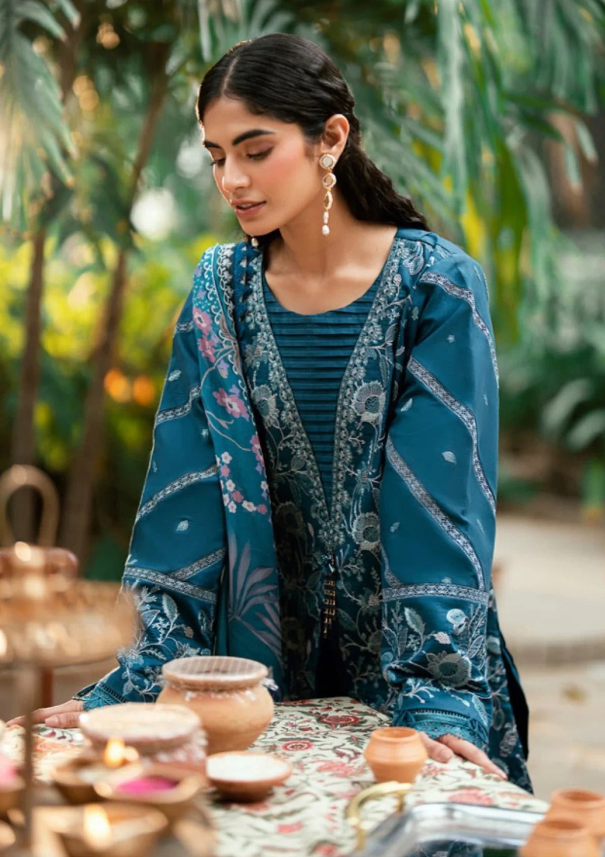 A woman wearing a teal blue traditional embroidered outfit with floral patterns, standing at a table with clay pots in a garden setting.
