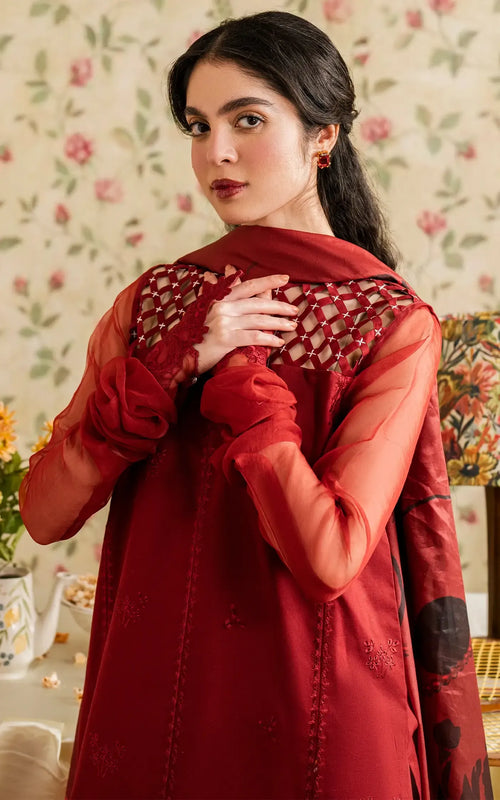 Woman wearing a red traditional outfit with intricate designs, standing against a floral wallpapered wall.