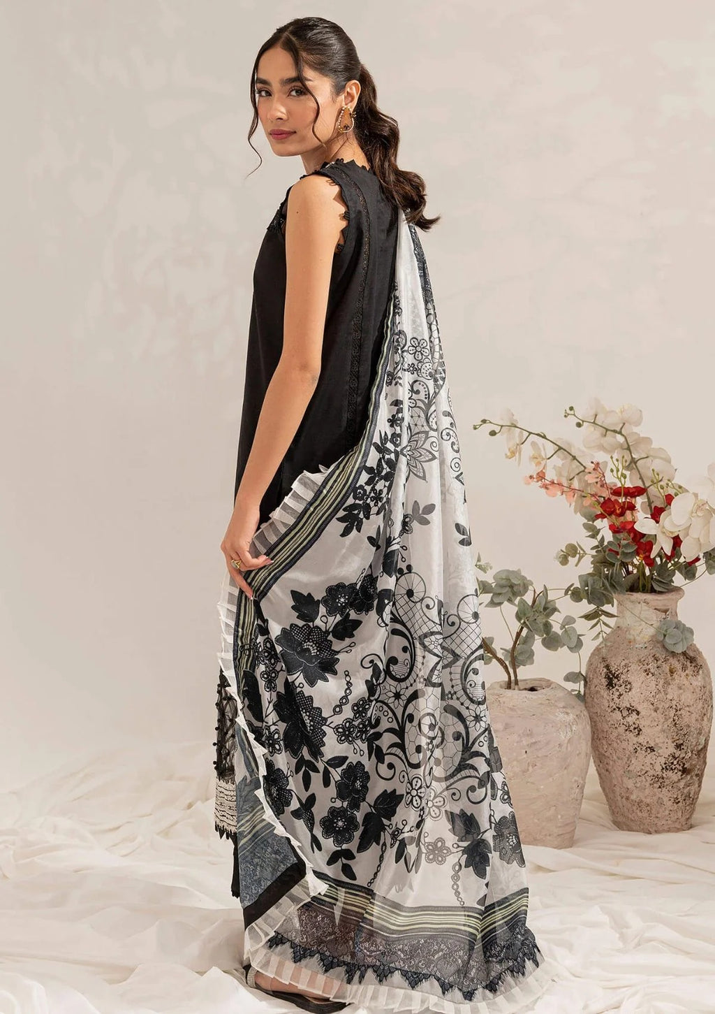 A woman models a black and white printed saree with a black sleeveless top, standing in a studio setting with decorative vases in the background.