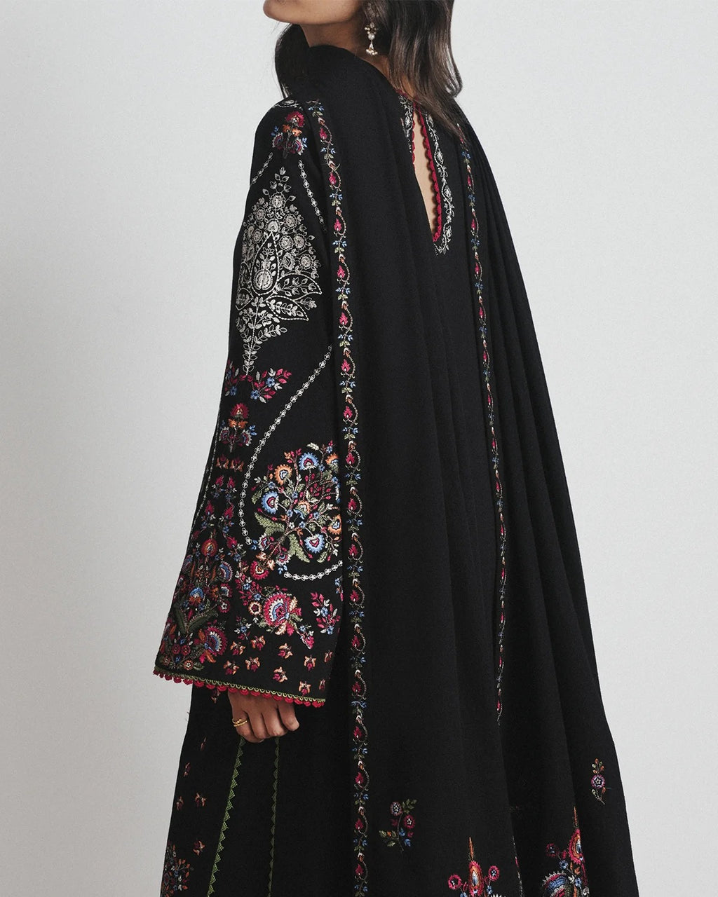 A woman wearing a black traditional embroidered outfit with ornate multicolor floral embroidery on the sleeve and border, shown from a side-back angle against a light gray studio background.