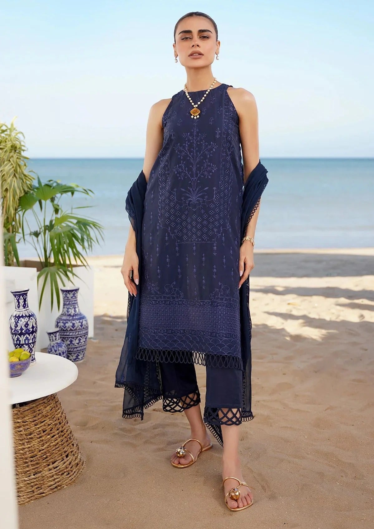 A woman stands on a sandy beach wearing a navy blue embroidered kurta with matching draped sleeves and cropped pants. The background shows the ocean, sky, and beachside decor.