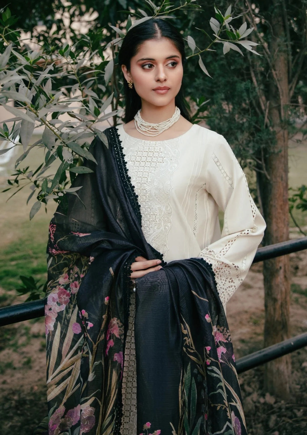 A woman wearing a cream-colored embroidered kurta with a black floral dupatta, standing outdoors in front of green trees.