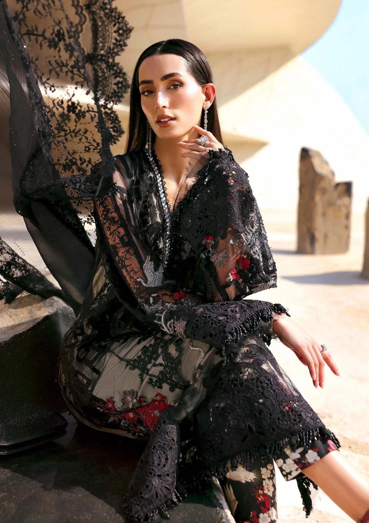 A woman seated outdoors in a desert-like architectural setting, wearing a sheer black lace robe with floral embroidery and fringe details over a patterned garment.