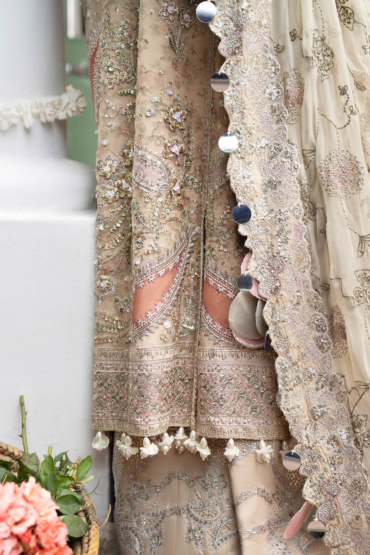 Close-up of an ornate traditional embroidered garment in warm beige and blush tones, with intricate metallic embroidery, tassel trim, and decorative buttons, shown hanging beside a white architectural column.