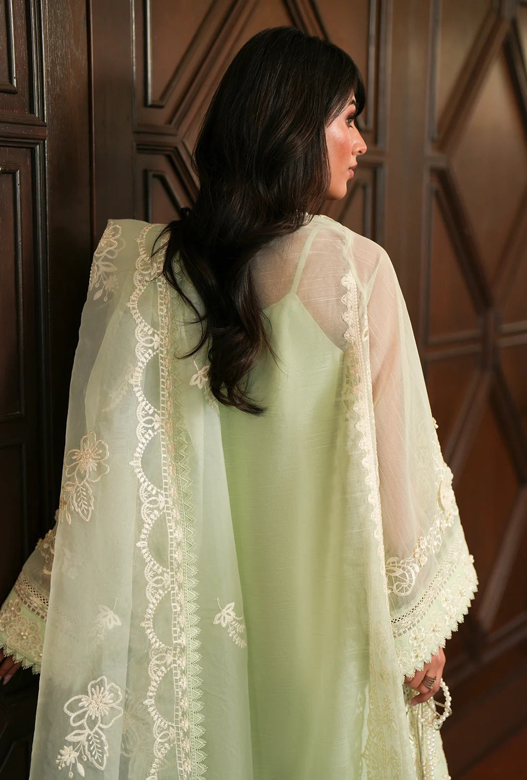 A woman shown from the back wearing a pale green dress with a sheer white embroidered shawl draped over her shoulders, posed in front of a dark wood paneled door.