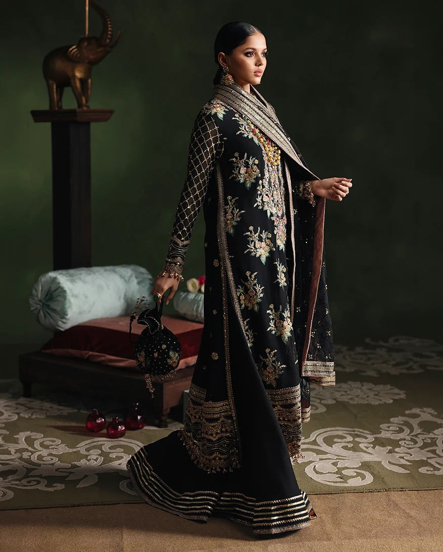 A woman models a black traditional embroidered outfit with gold and beige floral detailing, a matching draped scarf, and ornate metallic borders, posed in an indoor styled setting with a dark green wall and decorative rug.