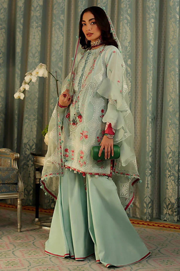 A woman models a traditional light mint green three-piece salwar kameez suit with floral embroidery, wide-leg pants, and a matching sheer dupatta in an ornate room with patterned curtains.