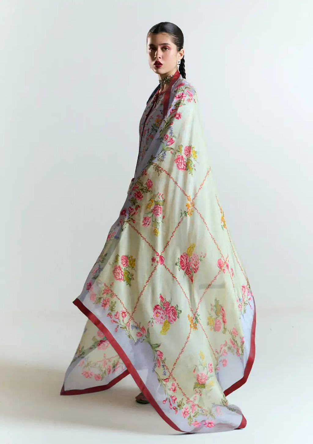 A woman models a long, flowing dupatta with a pale sage green base, decorated with pink and red floral motifs and a red border, set against a plain light gray studio background.