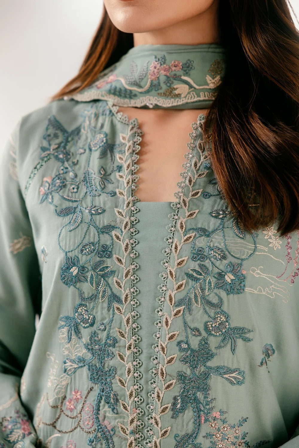 A close-up of a model wearing a sage green embroidered traditional blouse with intricate floral and vine patterns in blue, pink, and gold thread.