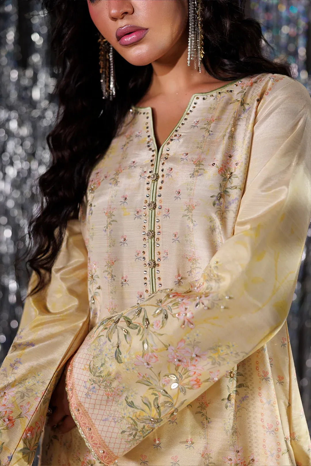 A close-up of a woman wearing a beige satin kurta with delicate floral embroidery in pink, green, and yellow, featuring a split neckline with gold-tone studs and long bell sleeves.